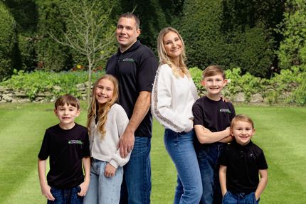 Five smiling people standing together in a green park, with trees and grass behind them
