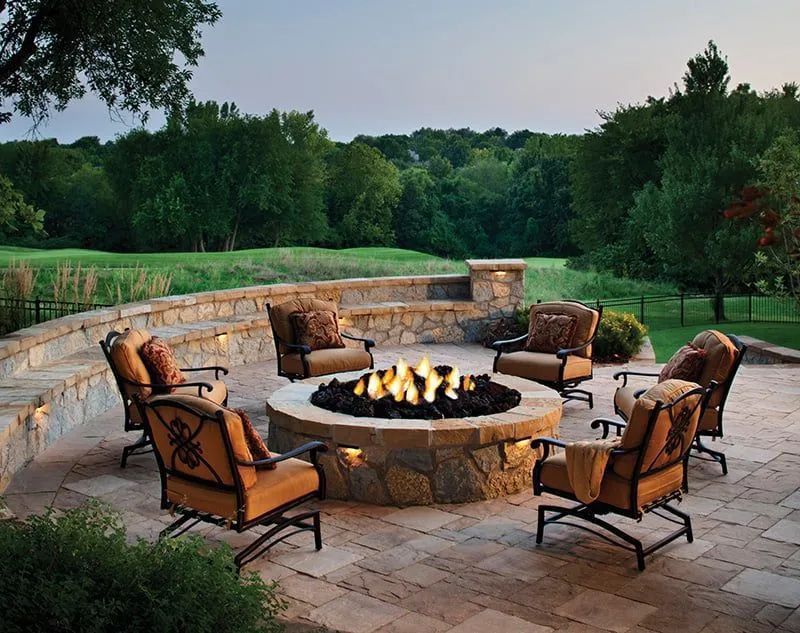 An outdoor stone patio with a fire pit surrounded by five cushioned armchairs in a scenic, grassy landscape at dusk.