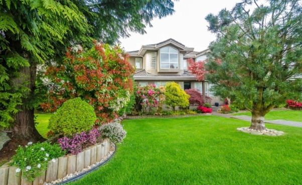 A two-story suburban house viewed from a front yard featuring a manicured lawn and various shrubs and trees.