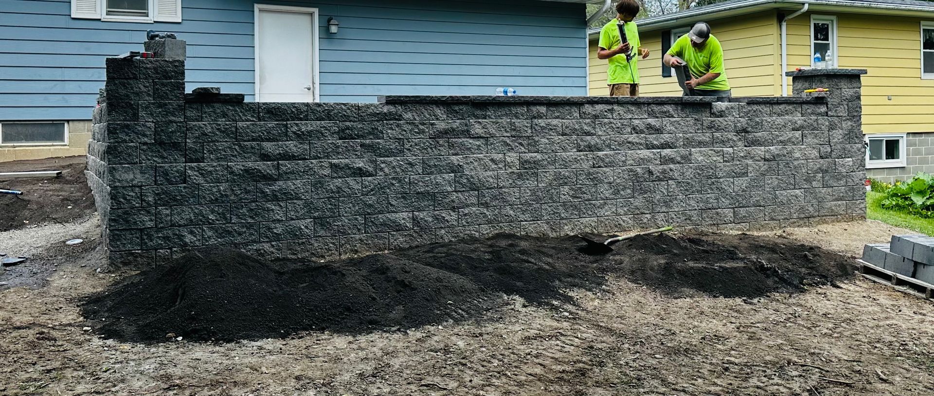 Stone retaining wall under construction in front of a house, with two workers in safety vests.