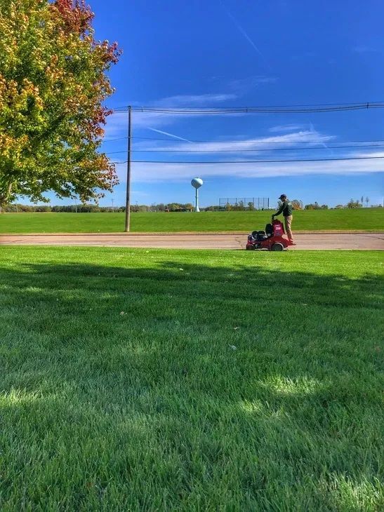 A person riding a red lawn mower on a grass field under a bright blue sky with a water tower in the distance.