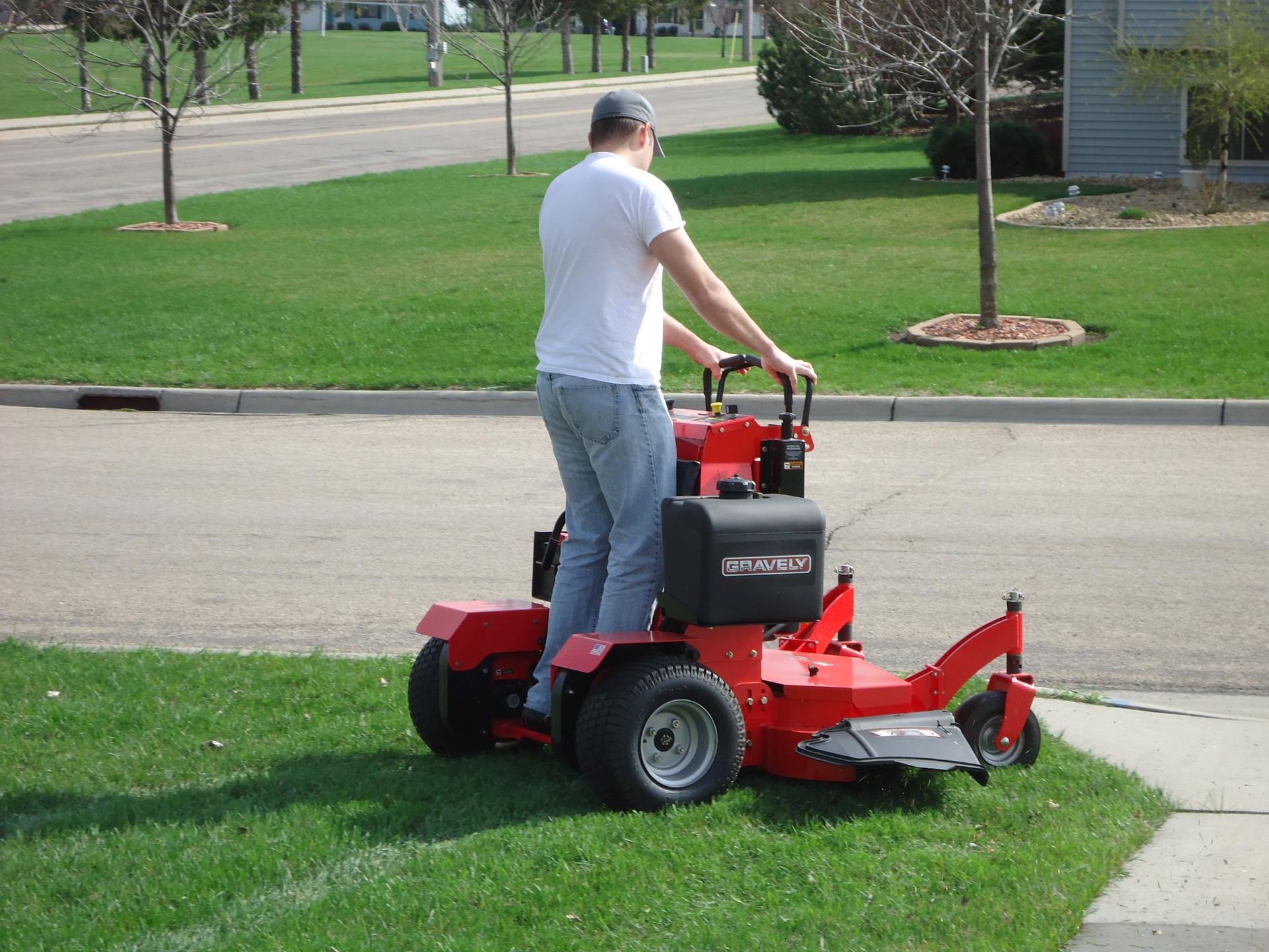 A person operates a red stand-on mower while driving onto a grass lawn from a paved road in a residential area.