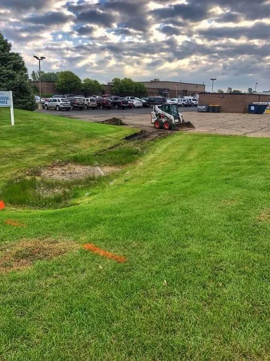 A white skid steer loader operates on a muddy path between a parking lot and a grassy lawn under a cloudy sky.
