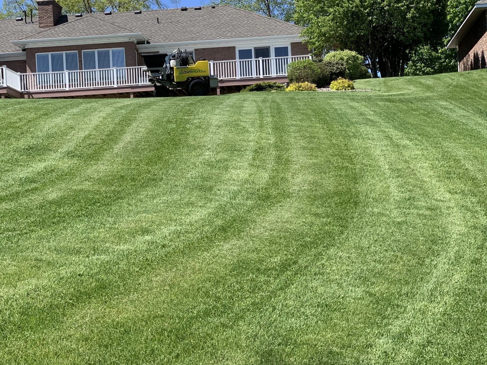 A yellow lawn mower on a steep, freshly mowed grassy hill in front of a residential house with a large deck.