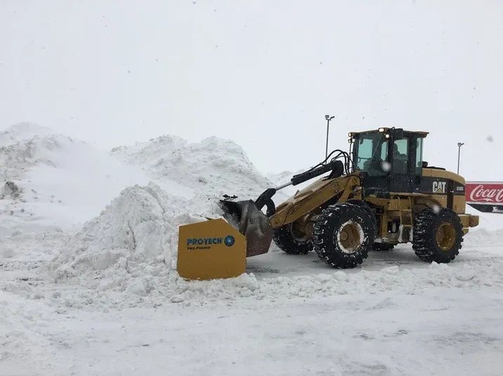 A yellow Caterpillar front-end loader with a snow pusher attachment clearing a large pile of snow in a snowy lot.