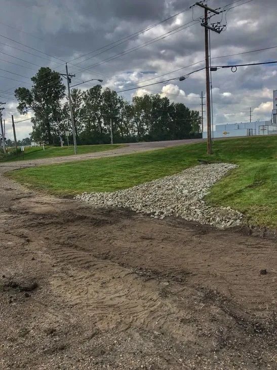 A gravel drainage channel cut into a green grassy slope beside a dirt road under a cloudy sky.