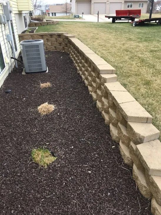 A retaining wall separates a mulched garden bed, containing a central AC unit and three small plants, from a lawn.