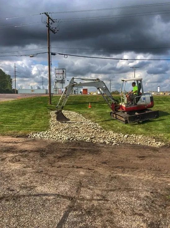 A construction worker operates a small excavator to place a gravel drainage area on a grassy roadside under a cloudy sky.