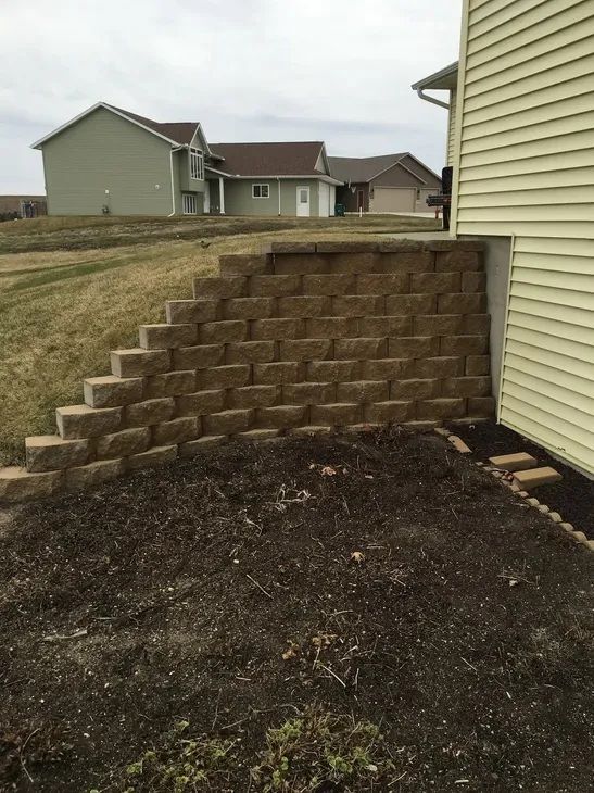 A stone retaining wall built along the side of a house with dirt ground in the foreground and neighboring houses behind.
