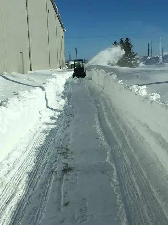 A piece of heavy machinery clears a path between a tall wall and a high pile of snow under a bright blue sky.