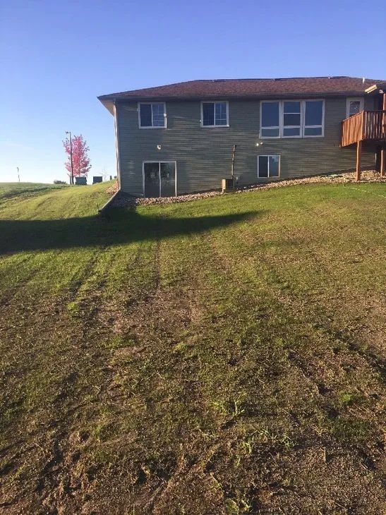 A light-colored split-level house sits on a grassy, sloped hill under a clear blue sky.