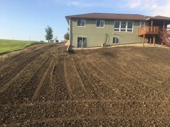 A house with a wooden deck overlooks a freshly tilled, sloping backyard under a clear blue sky.