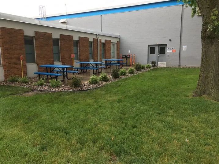 Blue picnic tables on a stone patio outside a building with brick accents, next to a large grassy lawn and a tree.