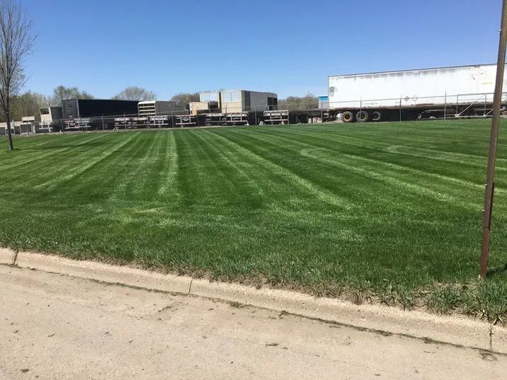 A mowed green lawn with neat grass stripes sits in front of industrial buildings and semi-truck trailers.