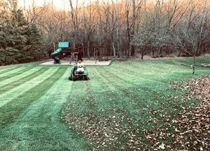 A person on a riding lawnmower cuts stripes into a grassy yard near a playground and forest edge at sunset.
