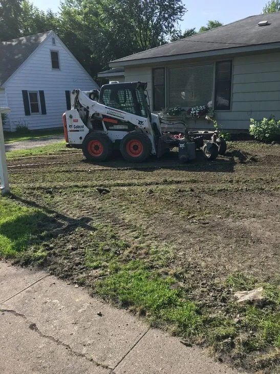 A white Bobcat skid-steer loader sits on a dirt-covered residential lawn in front of a house on a sunny day.
