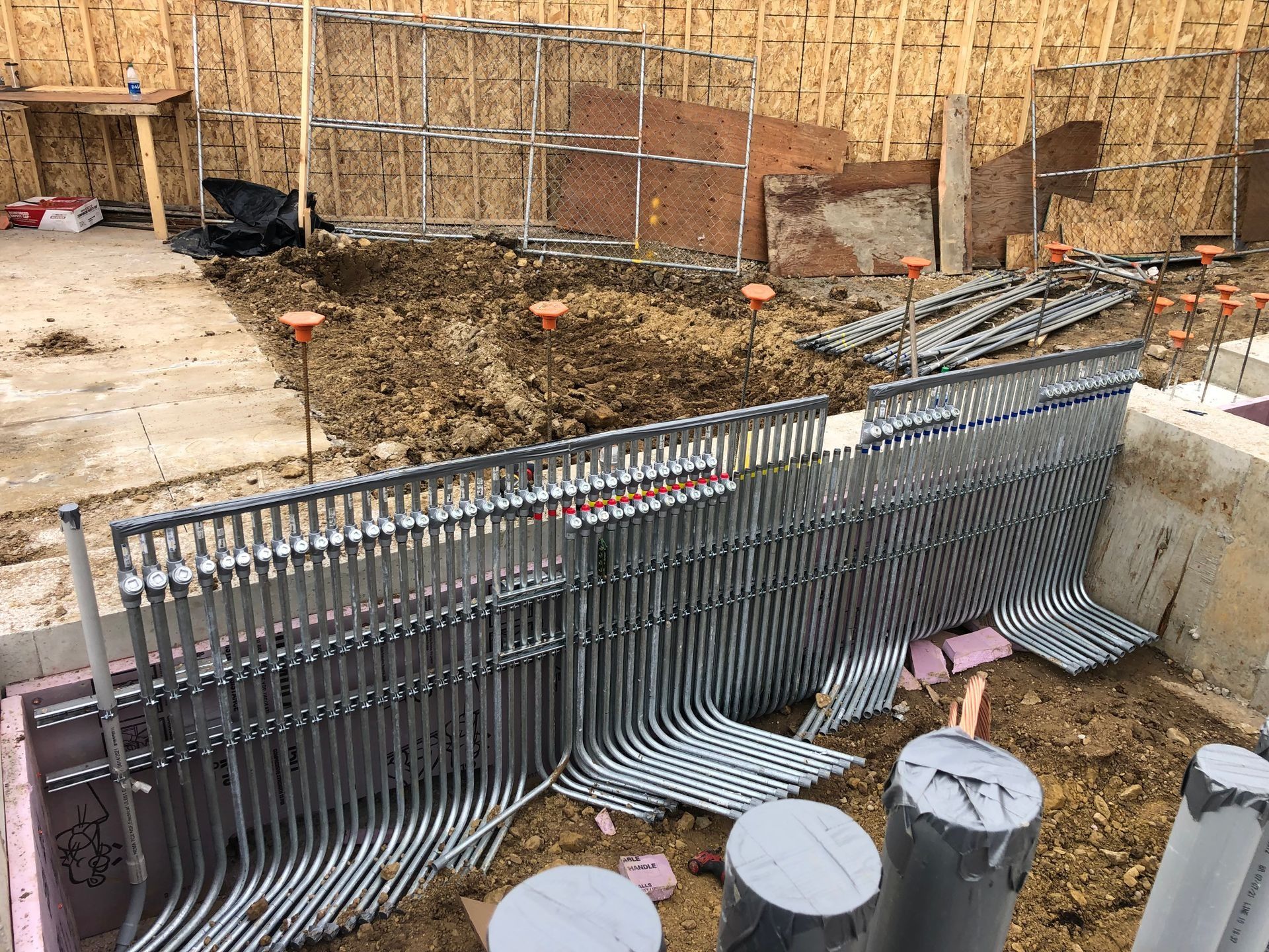 Construction site: Metal conduit pipes installed, emerging from a foundation trench; dirt, wood, and pink insulation visible.