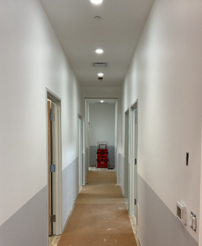 Long hallway with white and gray walls, recessed lighting, and closed doors. Red cart at the end.