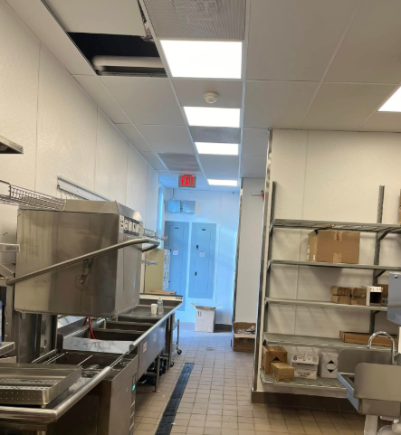Kitchen interior with stainless steel appliances, white walls, fluorescent lighting, and a partially open ceiling panel.