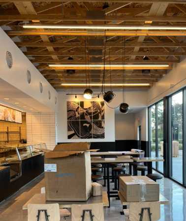 Interior of a restaurant under construction with exposed wood ceiling, tables, and large windows.