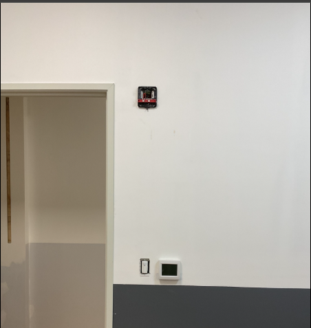 White wall with an open doorway, a fire alarm, a light switch, and a thermostat. The bottom of the wall is gray.