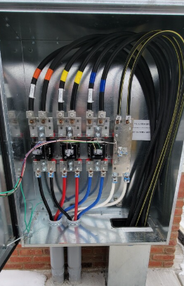 Electrical wiring in a metal box with labeled wires of various colors, set against a brick wall.