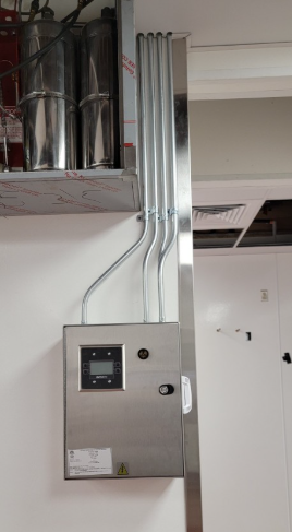 Stainless steel control panel with screen, mounted on a white wall with conduit and a stainless steel support.