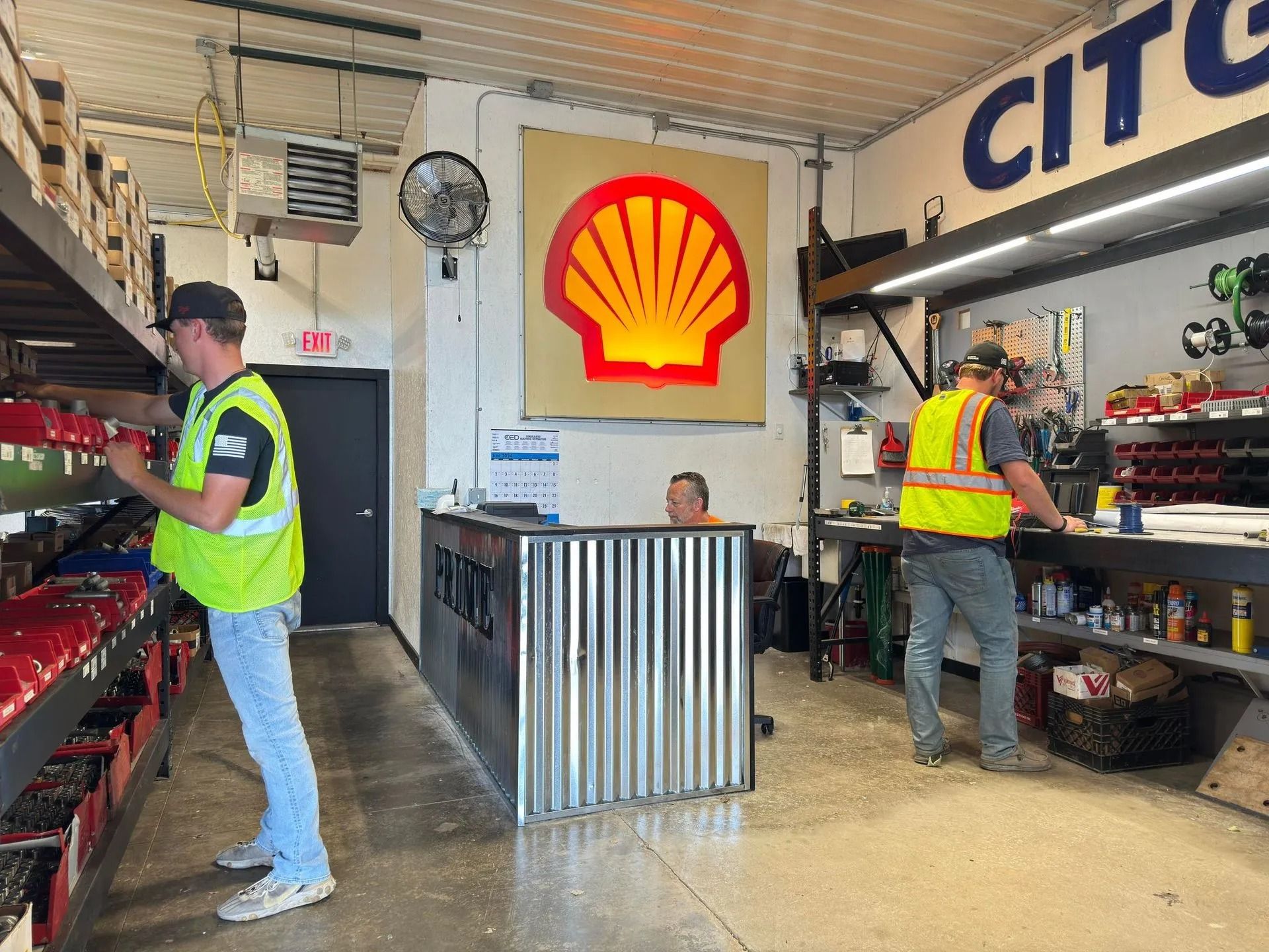 Two workers in a parts room, one looking at shelves, the other at a counter, both wearing safety vests, Shell logo in background.