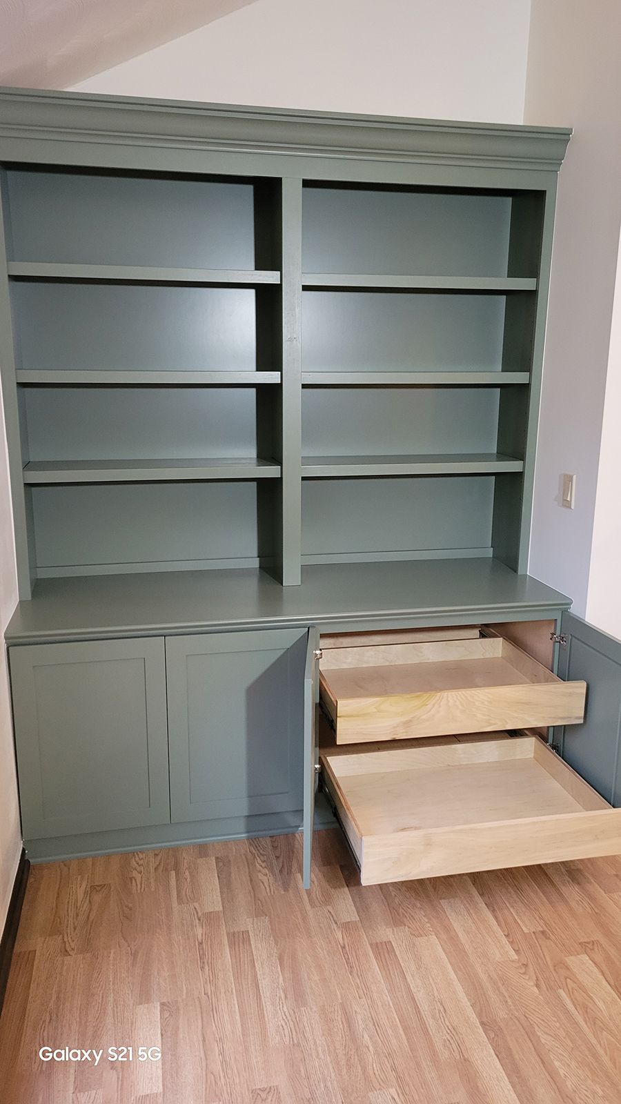 Built-in green cabinet with bookshelves, doors, and two open drawers on a wood floor.