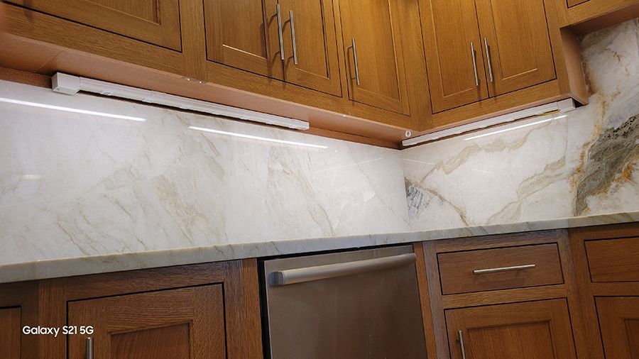 Kitchen cabinets with under-cabinet lighting and marble backsplash. Stainless steel dishwasher. Wooden cabinets.