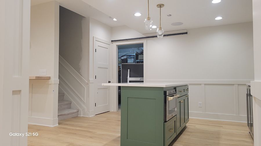 Green kitchen island in front of a white wall with wainscoting; light wood floors, stairs, and a door visible.