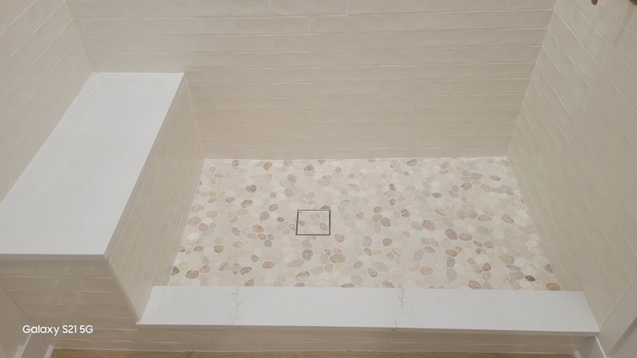 Tiled shower with pebble floor, built-in bench. Beige and white tones, a central drain.