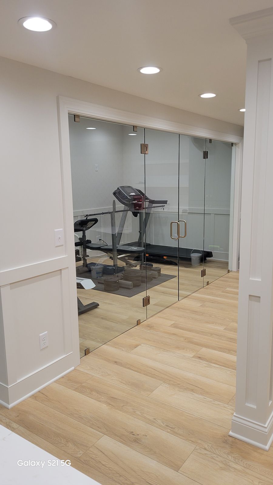 A home gym with a treadmill and exercise bike visible through a glass wall. Light wood floors and white walls.