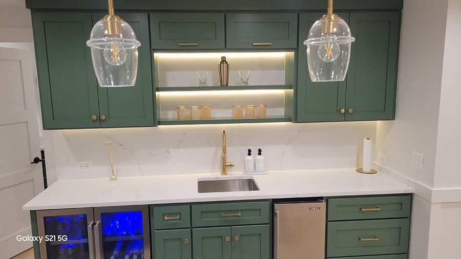 Green home bar with overhead cabinets, gold accents, white countertop, and sink.