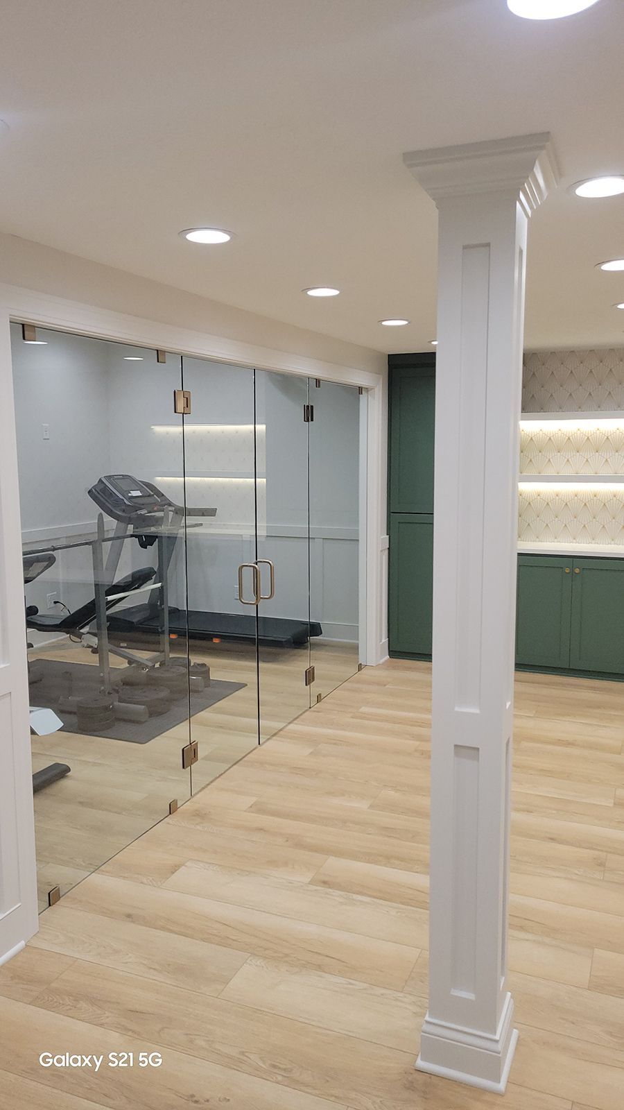 Home gym with glass walls, treadmill, and white column; light wood flooring and green cabinets.