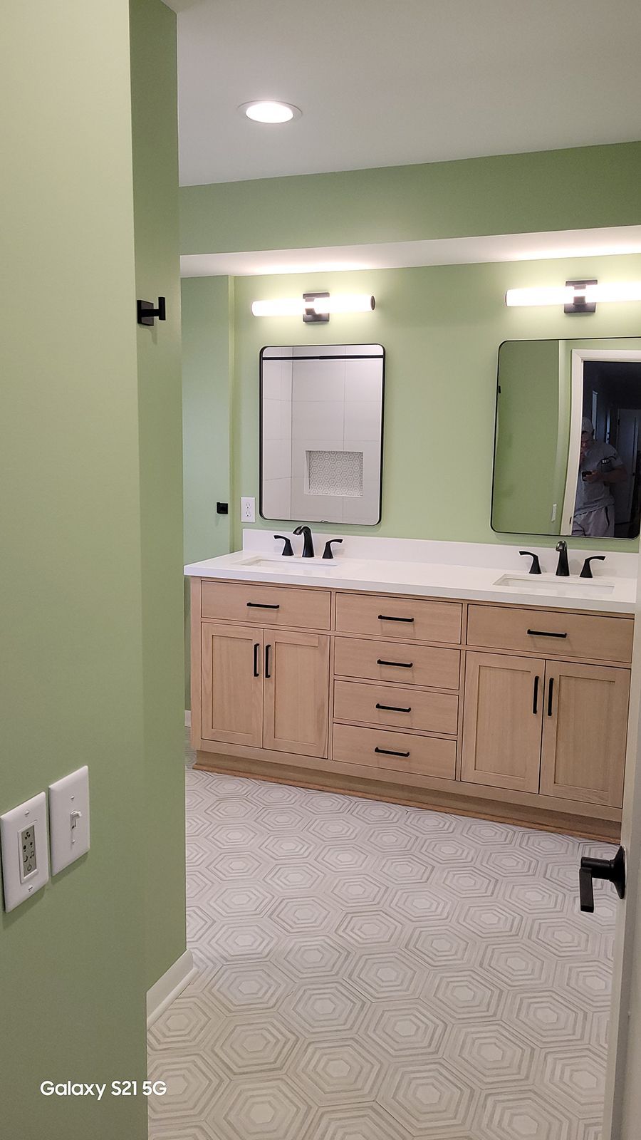 Bathroom with light wood cabinets, white countertops, black fixtures, and green walls.