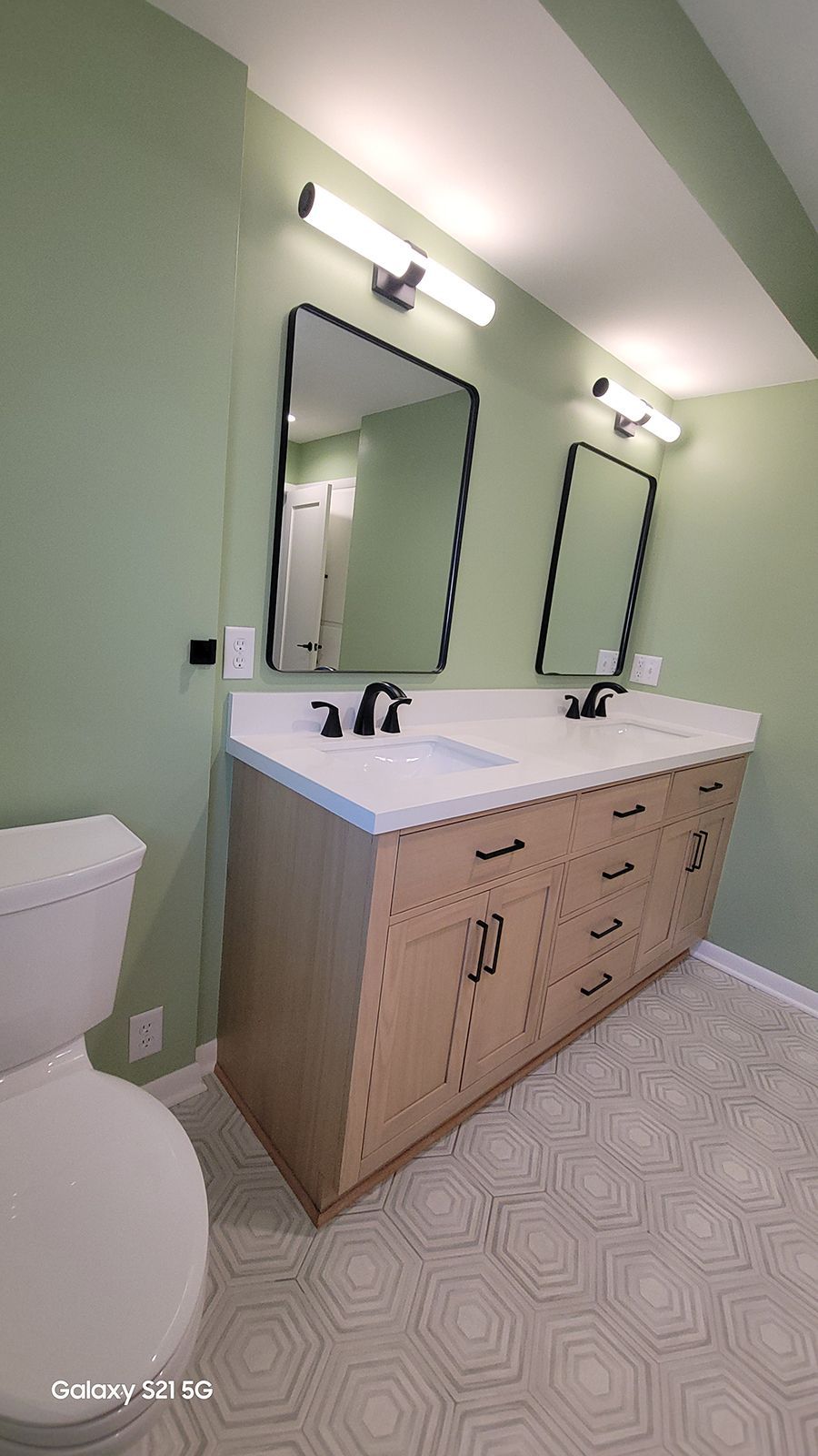 Bathroom with sage green walls, light wood vanity, black fixtures, and geometric floor tiles.