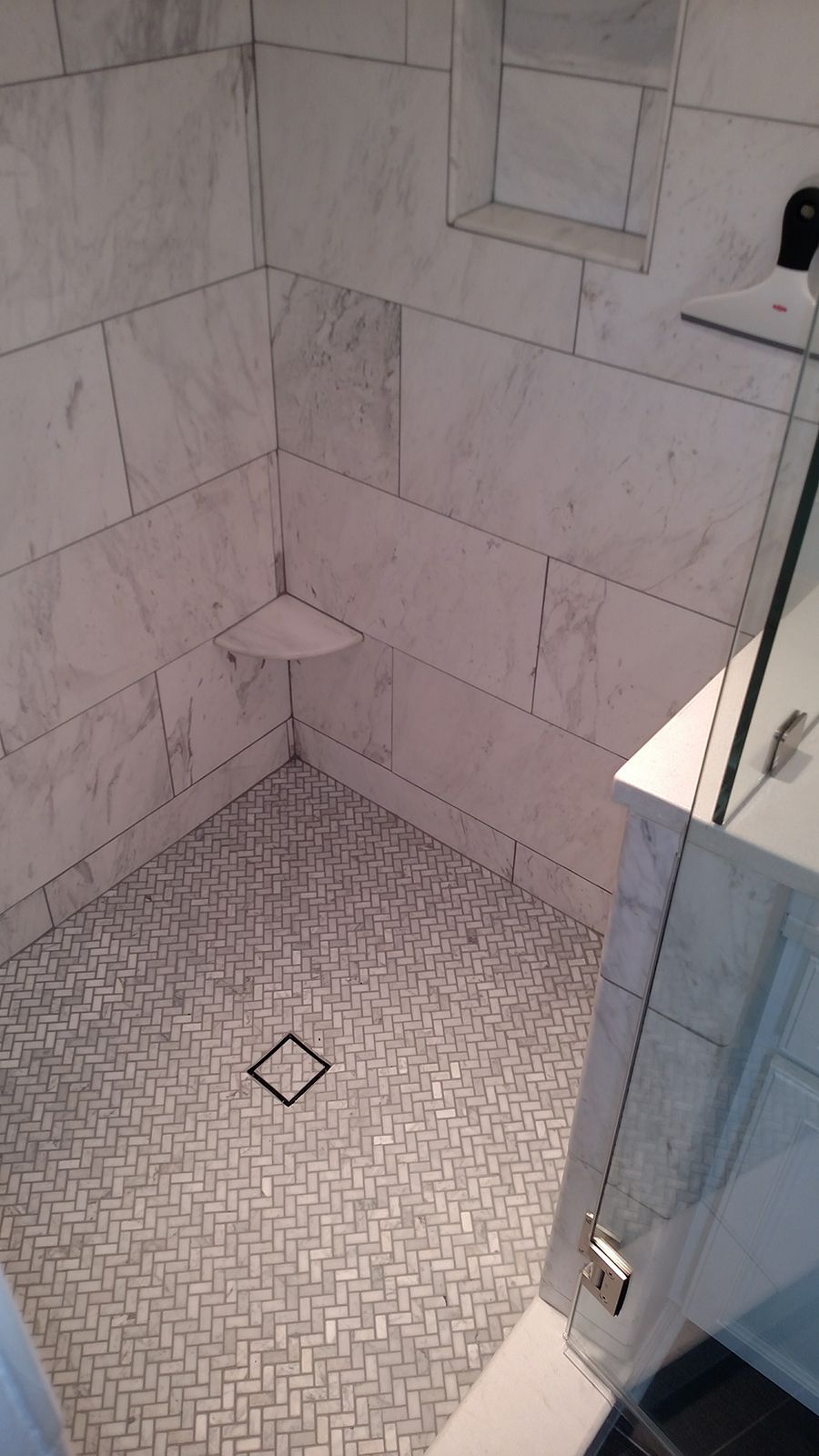 Tile shower with marble walls, herringbone floor, a built-in corner seat, and a glass door.
