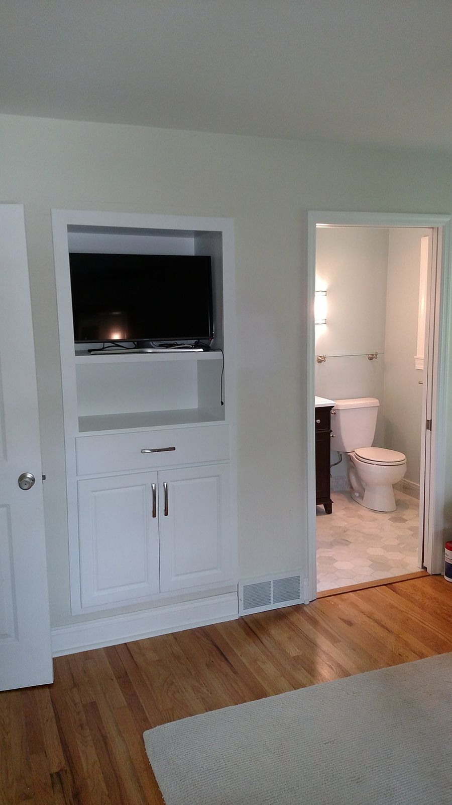 White built-in cabinet with TV, open doorway to bathroom with toilet, light-colored wood floor.