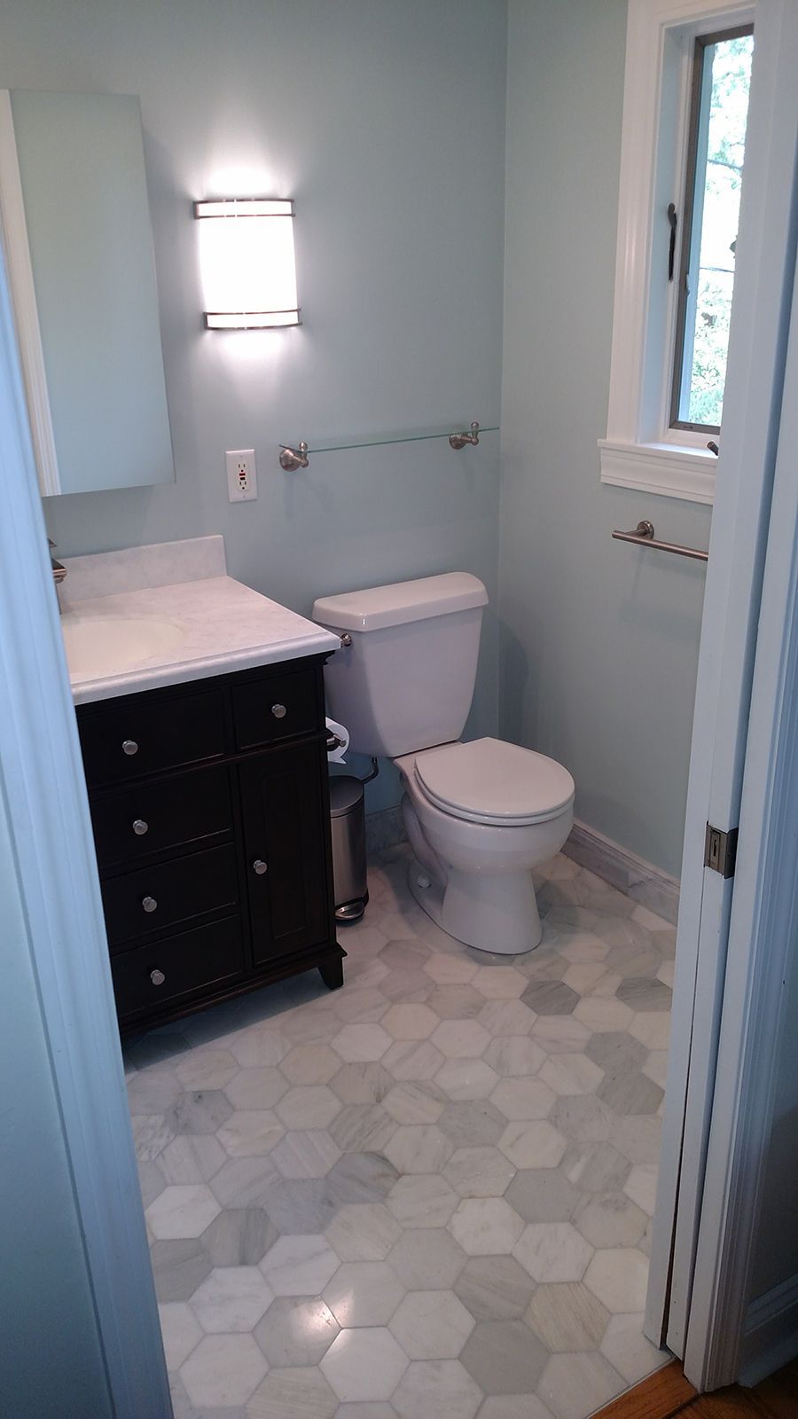 Small bathroom with light blue walls, hexagon tile floor, black vanity, and white toilet.
