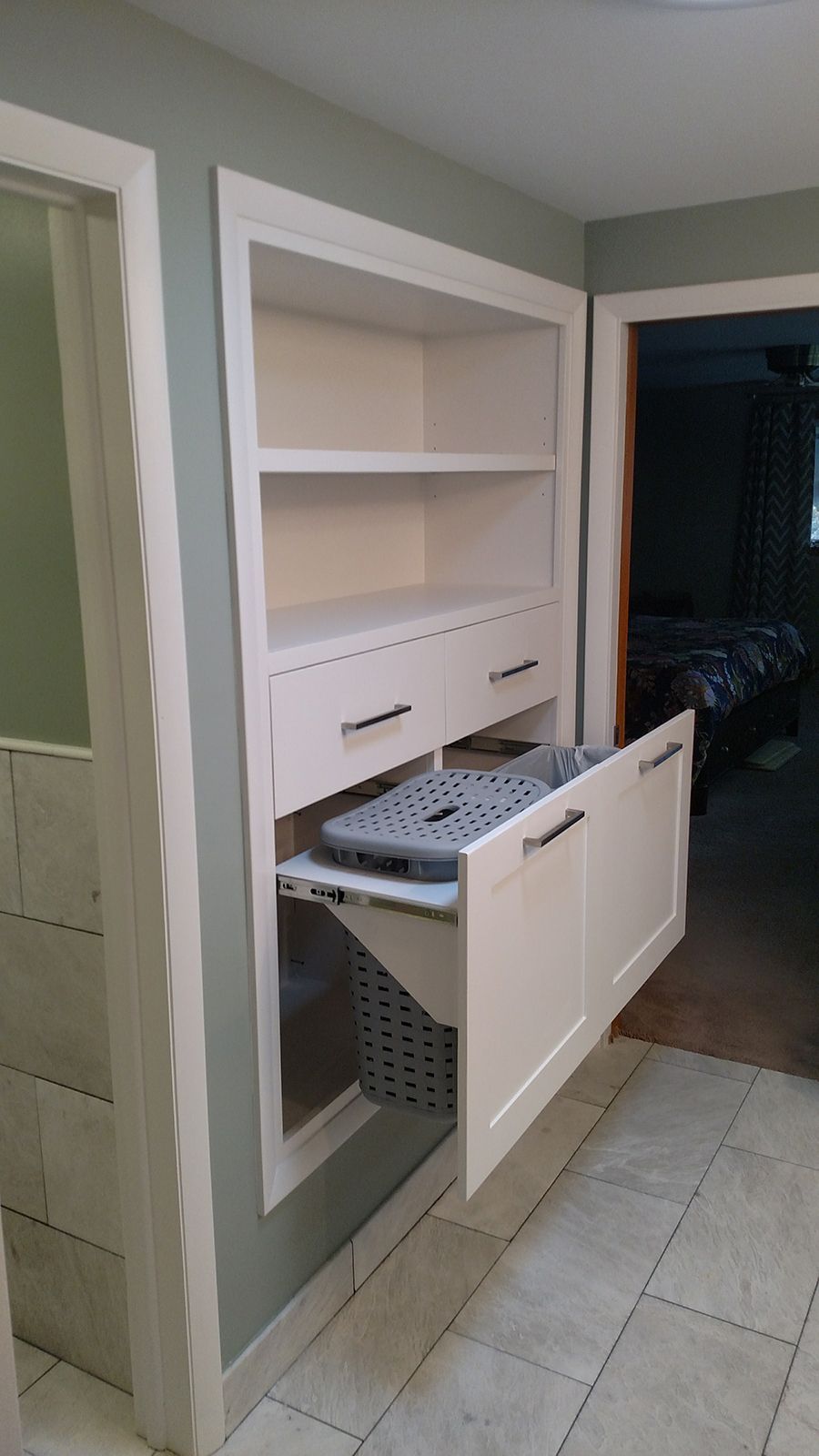 Built-in white laundry cabinet with shelves, drawers, and pull-out hamper in a bathroom nook.