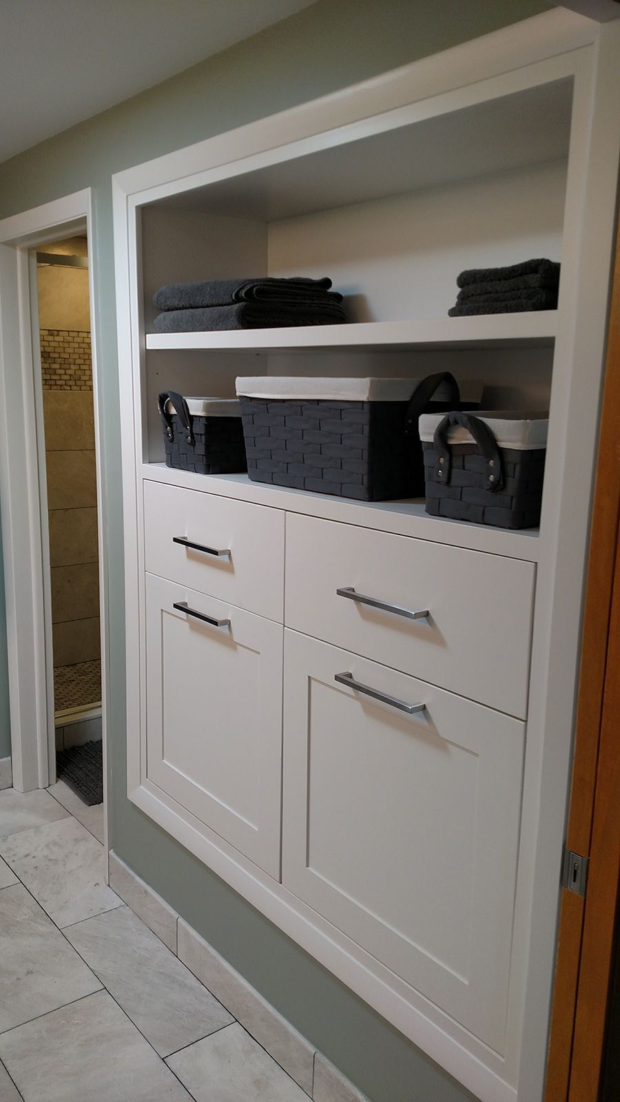 White built-in bathroom cabinet with shelves and drawers. Gray towels and storage baskets are displayed.