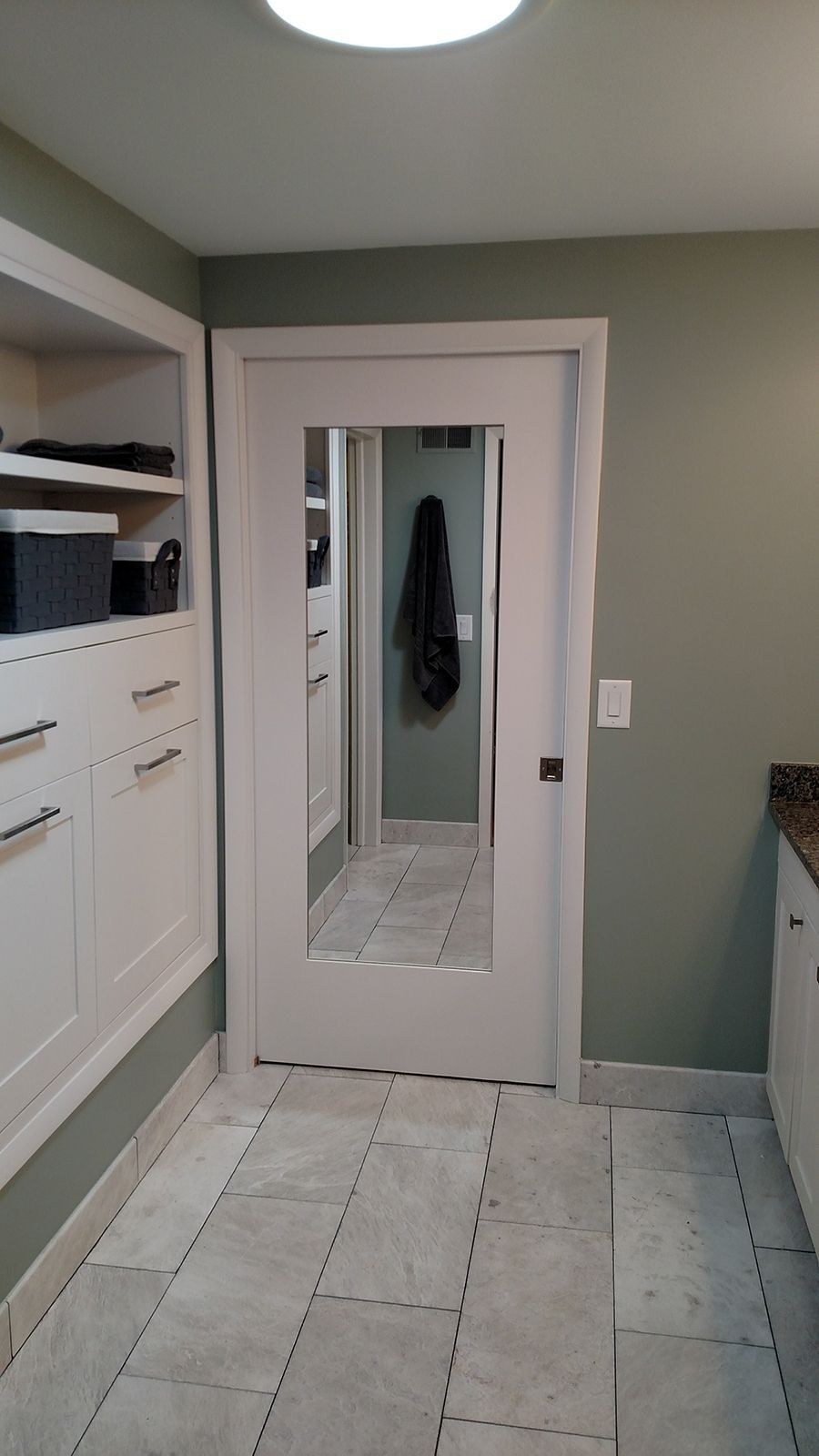 Bathroom doorway with a full-length glass panel; white trim, sage green walls, tiled floor, white cabinets.