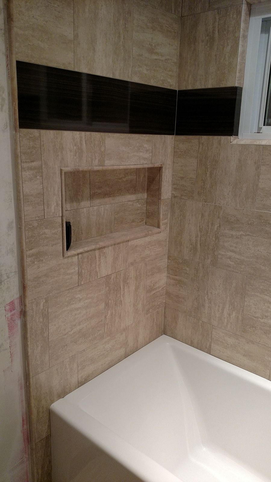 Bathroom with beige and black tiled walls, built-in shelf, and a white bathtub.