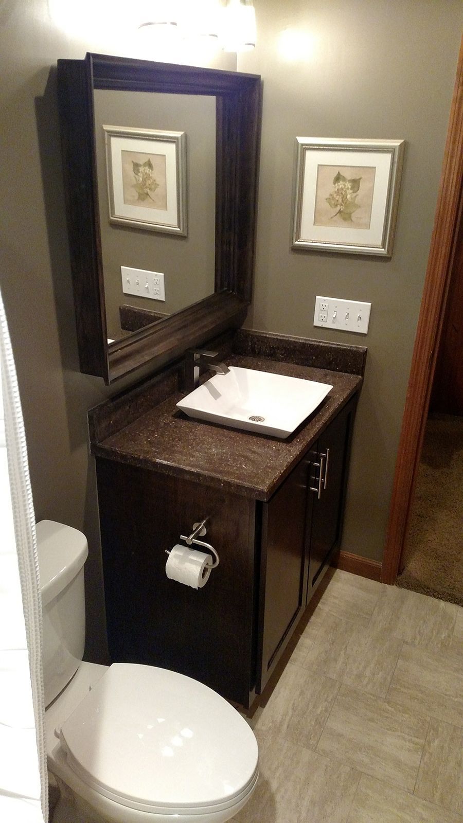 Small bathroom with dark brown vanity, mirror, toilet, and artwork on a gray wall.