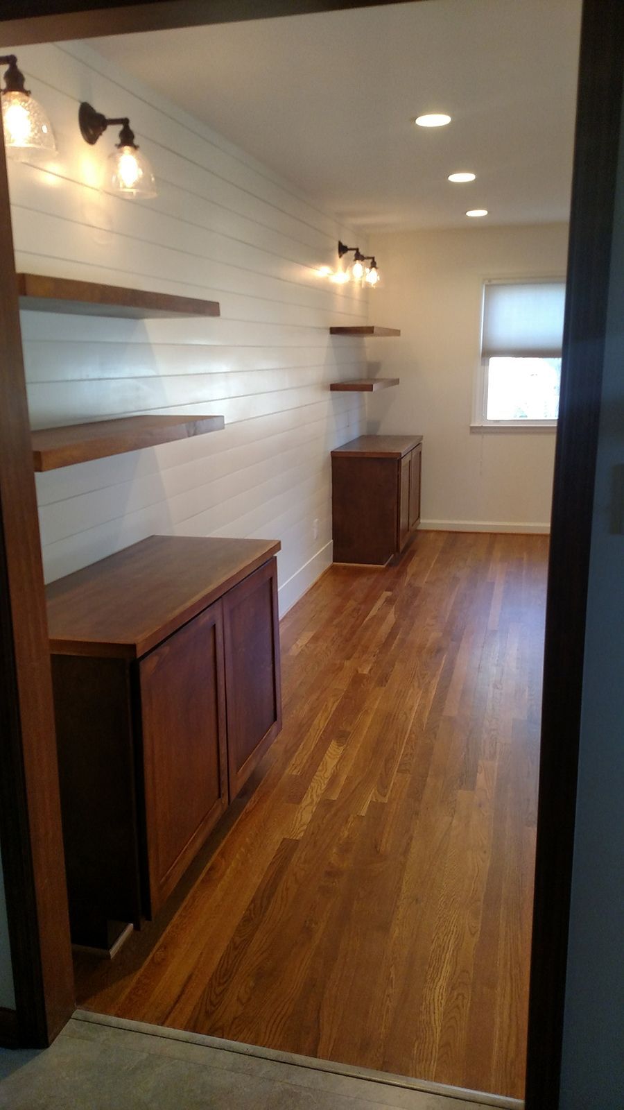 Wooden-floored room with white shiplap walls, wood cabinets, and floating shelves. Two sconces and recessed lighting.