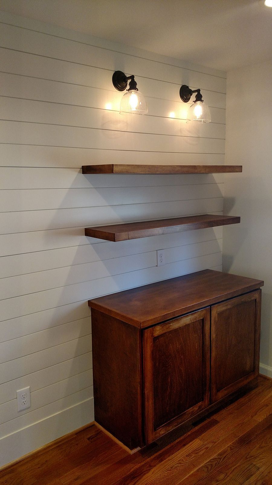 Wooden shelves and cabinet on a shiplap wall, illuminated by two sconces, over hardwood flooring.