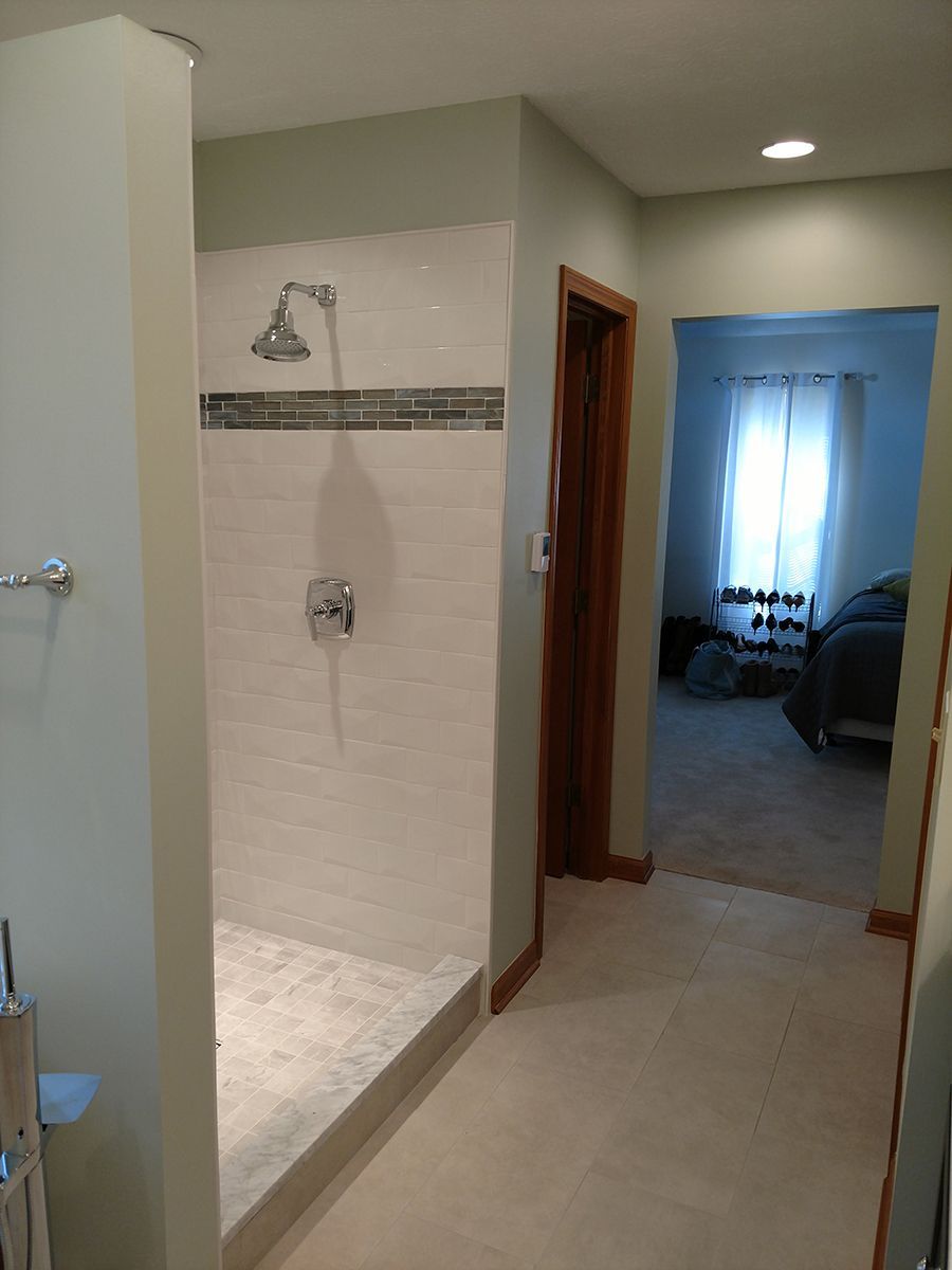 Bathroom with walk-in shower, white tile, and a view into a bedroom with a bed and window.
