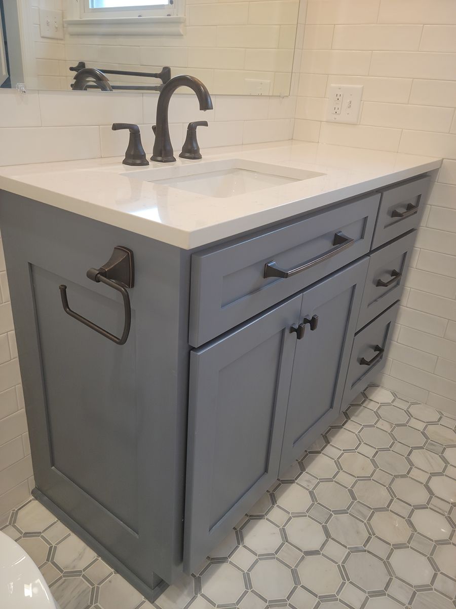 Gray bathroom vanity with white countertop, chrome faucet and hardware, set against white tile.