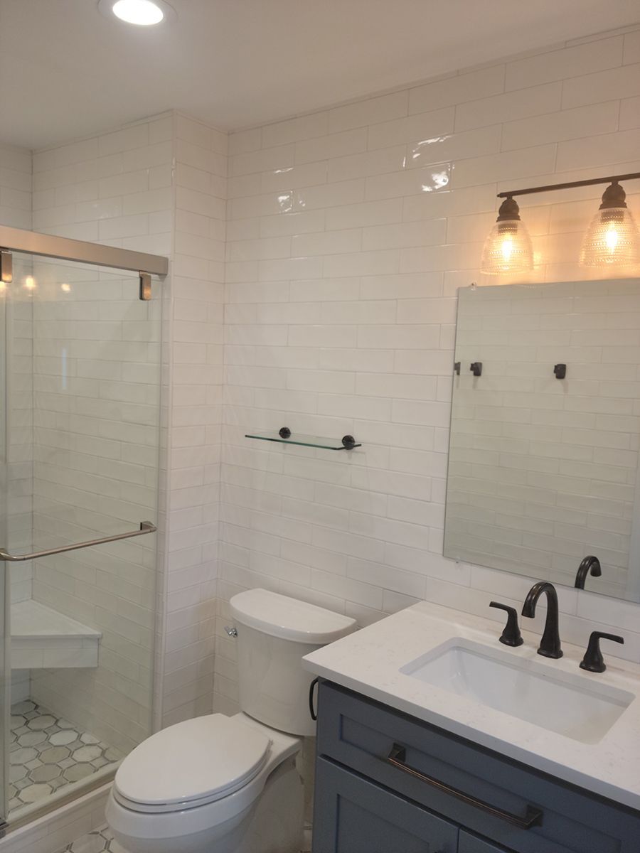 Modern bathroom with white tiled walls, glass shower, toilet, blue vanity, and a mirror.